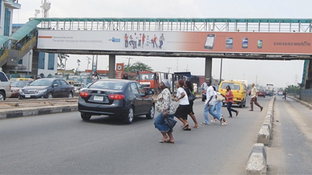 People crossing street in traffic with a pedestrian bridge visible in background.